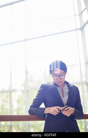 Attractive Asian entrepreneur in eyeglasses holding smartphone in hands while standing at spacious office lobby, glass wall on background Stock Photo