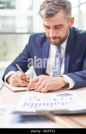Portrait of confident bearded financial manager wearing classical suit sitting at office desk and preparing annual accounts, blurred background Stock Photo