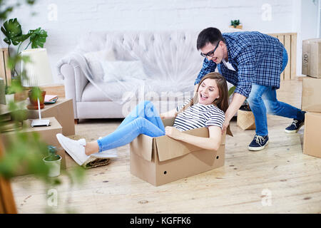 Lovely young couple having fun while riding in cardboard box at new apartment, interior design items and moving boxes on background Stock Photo