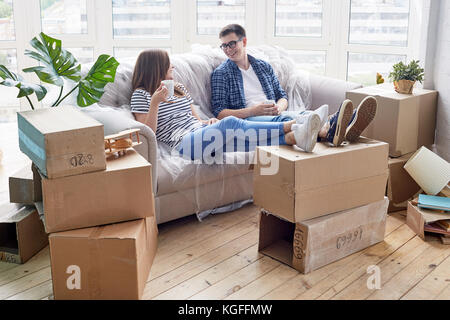 Cheerful young couple chatting animatedly with each other and drinking delicious cappuccino while taking short break from unpacking stuff, piles of mo Stock Photo