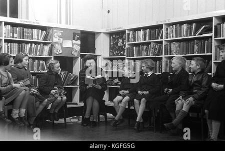 1955, historical picture shows a female teacher sitting and reading a story to a small group of young children in a corner of the school library. Stock Photo