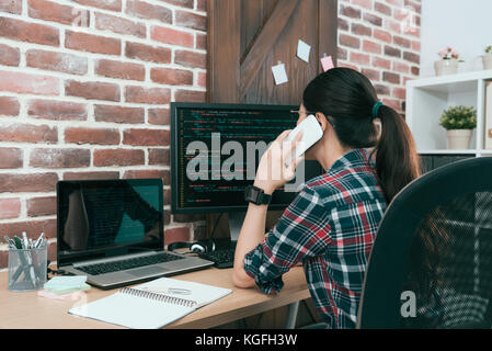 back view photo of pretty attractive female computer software development worker looking at code document and using mobile phone calling with client d Stock Photo