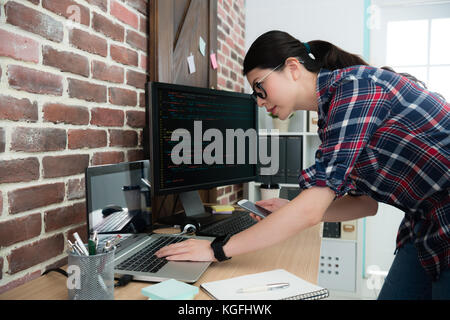 smiling elegant female programmer manager holding mobile smartphone standing in front of company computer and watching document checking website desig Stock Photo