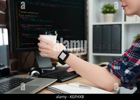smiling attractive female programmer wearing smartwatch with blank screen and holding hot coffee cup sitting in office using computer work. Stock Photo