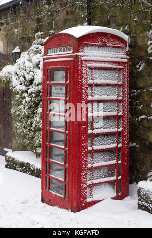 Cold, snowy winter village scene with close-up of iconic red telephone box & pavement covered in white snow - Hawksworth, West Yorkshire, England, UK. Stock Photo