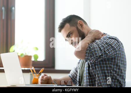 Troubled bearded man massages aching neck with a pained expression, working on laptop computer, deep in thoughts. Stock Photo