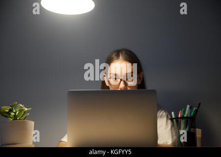 Overworked attentive young adult woman in glasses working on laptop computer at workplace desk late at night. Exam student preparing for test at home  Stock Photo