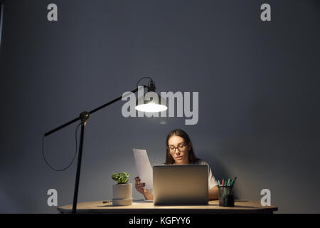Overworked tired young adult female accountant working on laptop computer, making report, checking contract order details late at night to meet deadli Stock Photo