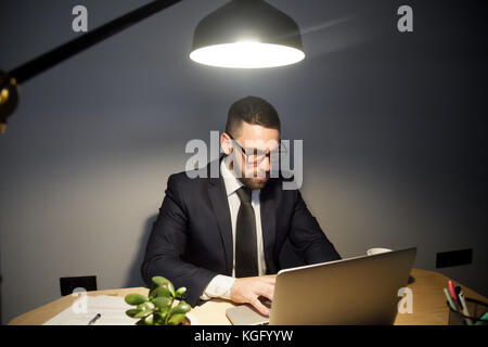 Attentive bearded man in glasses working on project late at night, reading a contract, email or difficult scientific article, typing messages, writhin Stock Photo