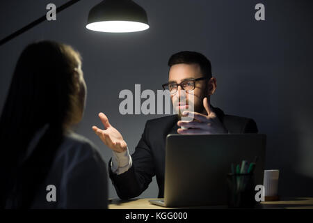 Two businesspeople communicating in late evening meeting in dark office, discussing problem with new project. Man gesturing, persuading business partn Stock Photo