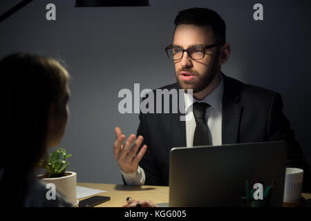 Male boss in suit explains to female employee project ideas and plans in late evening meeting still at workplace. Businesspeople working at night brie Stock Photo