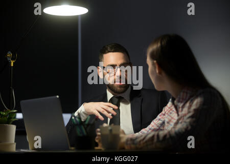 Two business people in late evening meeting discussing current projects. Bearded man in glasses and suit tells female subordinate about tasks for next Stock Photo
