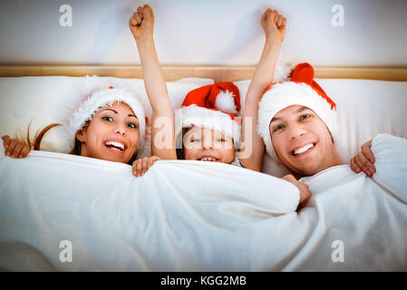 Smiling happy mom, dad and little girl in red hats lying in white bed covered by blanket and having fun. Stock Photo