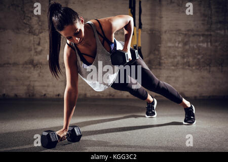 Young muscular woman doing plank exercise with dumbell at the gym. Stock Photo
