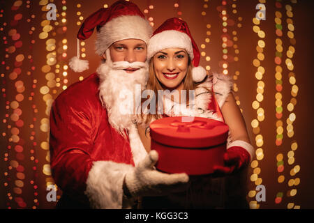 Beautiful young smiling couple in Santa Claus costume holding red present and looking at camera. Stock Photo