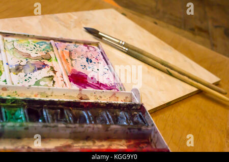 Dirty pallet and watercolor set with two wooden brushes on the table and a mug of water. Stock Photo