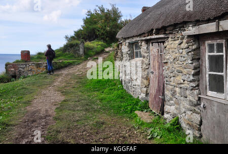 A walker on the coast path pauses to look at an old shellfish boiling pot, on the cliff overlooking the sea with old fishermens hut .Prussia cove UK Stock Photo