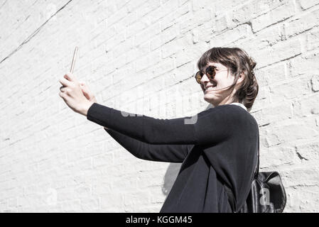 Young woman wearing sunglasses smiling and taking a selfie in front of a white brick wall as background Stock Photo