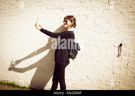 Young woman dressed in black with a small leather backpack wearing sunglasses smiling and taking a selfie in front of a white brick wall as background Stock Photo