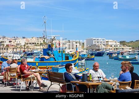 Tourists relaxing at a pavement cafes along the waterfront with traditional Maltese fishing boats in the harbour and views towards the town, Marsaxlok Stock Photo