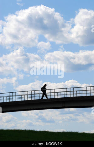 Millennial on the move, holding smartphone, crossing bridge. Silhouette of young man against clear blue sky. Stock Photo