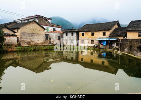 Traditional Chinese buildings are reflected in a pond. Behind a mountainous backdrop is wreathed in low cloud. Stock Photo