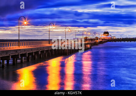 Stormy sunset at Port Philip bay around St Kilda beach historic timber Jetty with street light illumination reflecting in blue waters of the bay. Melb Stock Photo