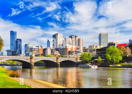 Princes bridge in Melbourne city across Yarra river on a sunny day in view of high-rise towers and modern urban architecture.