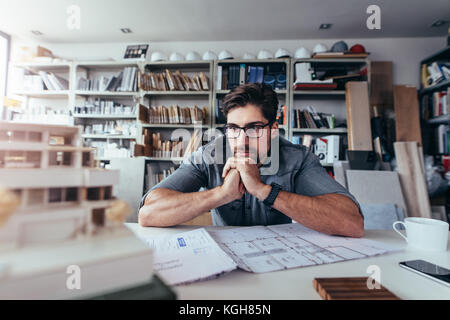Young architect looking at architecture house model and thinking about new ideas. Male designer sitting in office and working on new project. Stock Photo