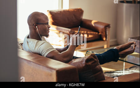 Man wearing earphones looking at mobile phone sitting on a sofa. Man listening to music and relaxing at home. Stock Photo