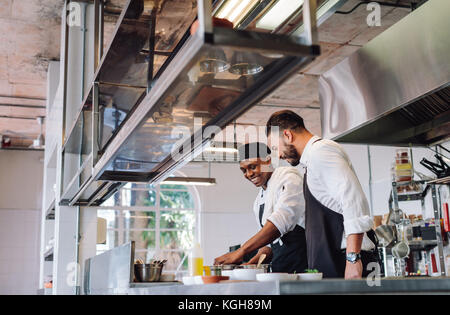 Happy male chefs cooking food at cafe kitchen and talking. Two cooks preparing food in restaurant kitchen. Stock Photo