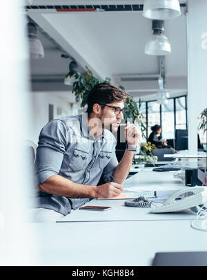 Vertical shot of thoughtful businessman looking at monitor. Young man sitting in office and thinking about work. Stock Photo