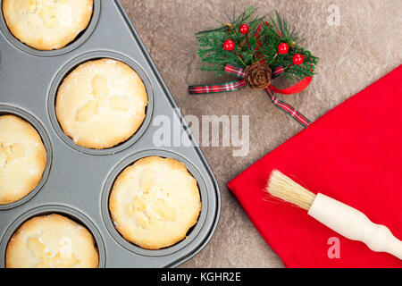 Fresh golden brown mince pies in a baking tray with a christmas motif and a pastry brush on a red napkin Stock Photo