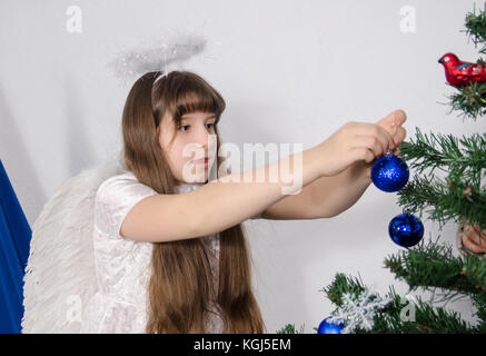 a girl in an angel costume decorates a Christmas tree with toys Stock Photo