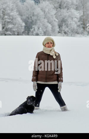 A girl in a light knitted hat and a dark winter jacket is standing next to a large black labrador dog among a field covered with snow. Stock Photo