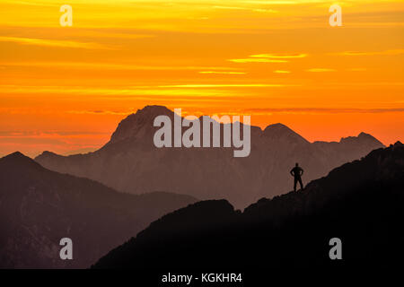 Happy successful winning man reaching mountain summit. Spectacular layered mountain ranges silhouettes with orange sunset sky. Stock Photo