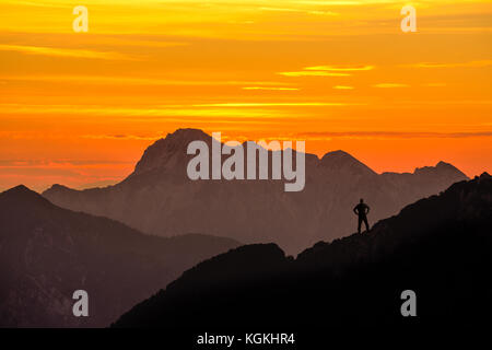 Happy successful winning man reaching mountain summit. Spectacular layered mountain ranges silhouettes with orange sunset sky. Stock Photo