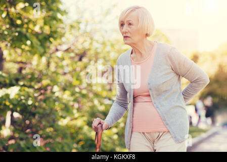 Unpleasant pain. Pleasant sad elderly woman leaning on the walking stick and holding her back while feeling pain there Stock Photo