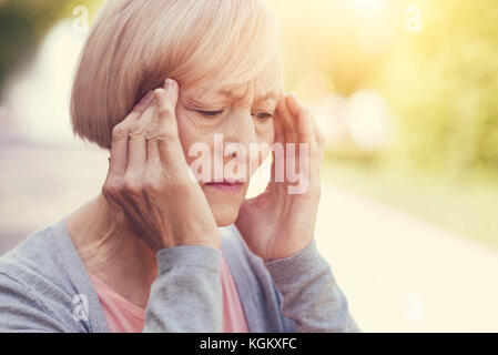 I feel headache. Cheerless elderly unhappy woman holding her temples and suffering from headache while being outdoors Stock Photo