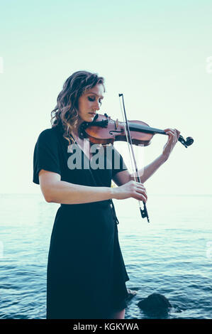 Beautiful young woman playing violin while standing by lake against clear sky Stock Photo