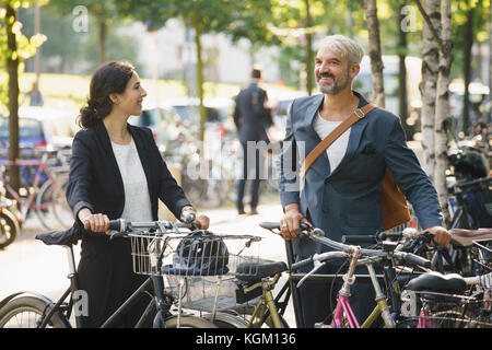 Smiling colleagues talking while standing with bicycles at parking lot Stock Photo