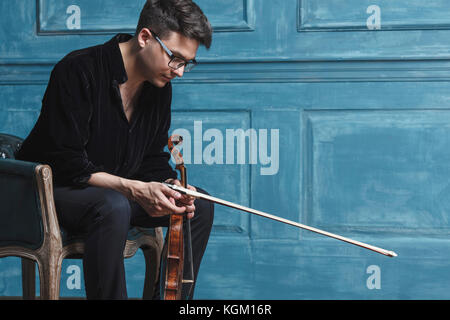 Young man holding violin while sitting on chair by blue wall Stock Photo