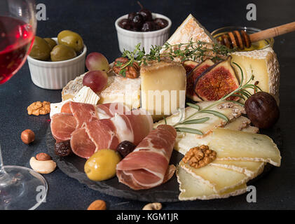 Glass of red wine and cheese plate with pieces moldy cheese, prosciutto, pickled plums, olives, grapes, figs, honey, pear, nuts on black slate backgro Stock Photo