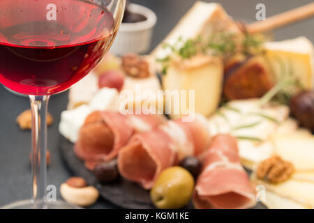 Glass of red wine and blur cheese plate with pieces moldy cheese, prosciutto, pickled plums, olives, grapes, figs, honey, pear, nuts on black slate ba Stock Photo