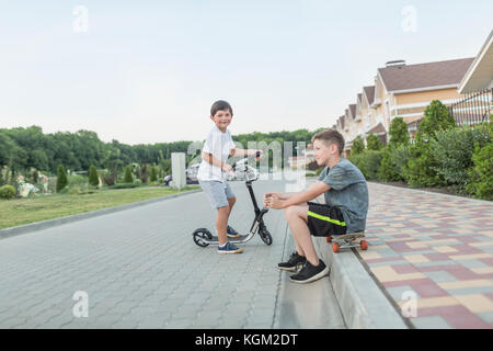 Boy sitting on skateboard while brother riding push scooter on cobbled street Stock Photo