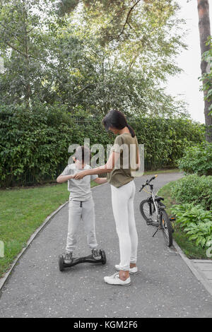 Mother assisting son in using self-balancing board on road against plants Stock Photo