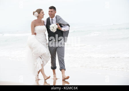 Happy bride and groom walking barefoot on the beach by the sea Stock Photo