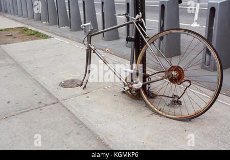 Broken, rusty bicycle with missing front wheel and dropped chain locked to a metal pole on a sidewalk Stock Photo