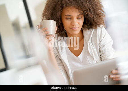 Mixed-race woman at home using tablet and drinking tea Stock Photo
