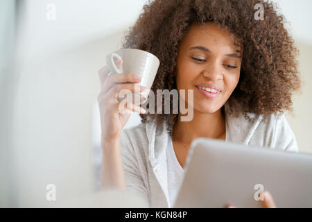 Mixed-race woman at home using tablet and drinking tea Stock Photo